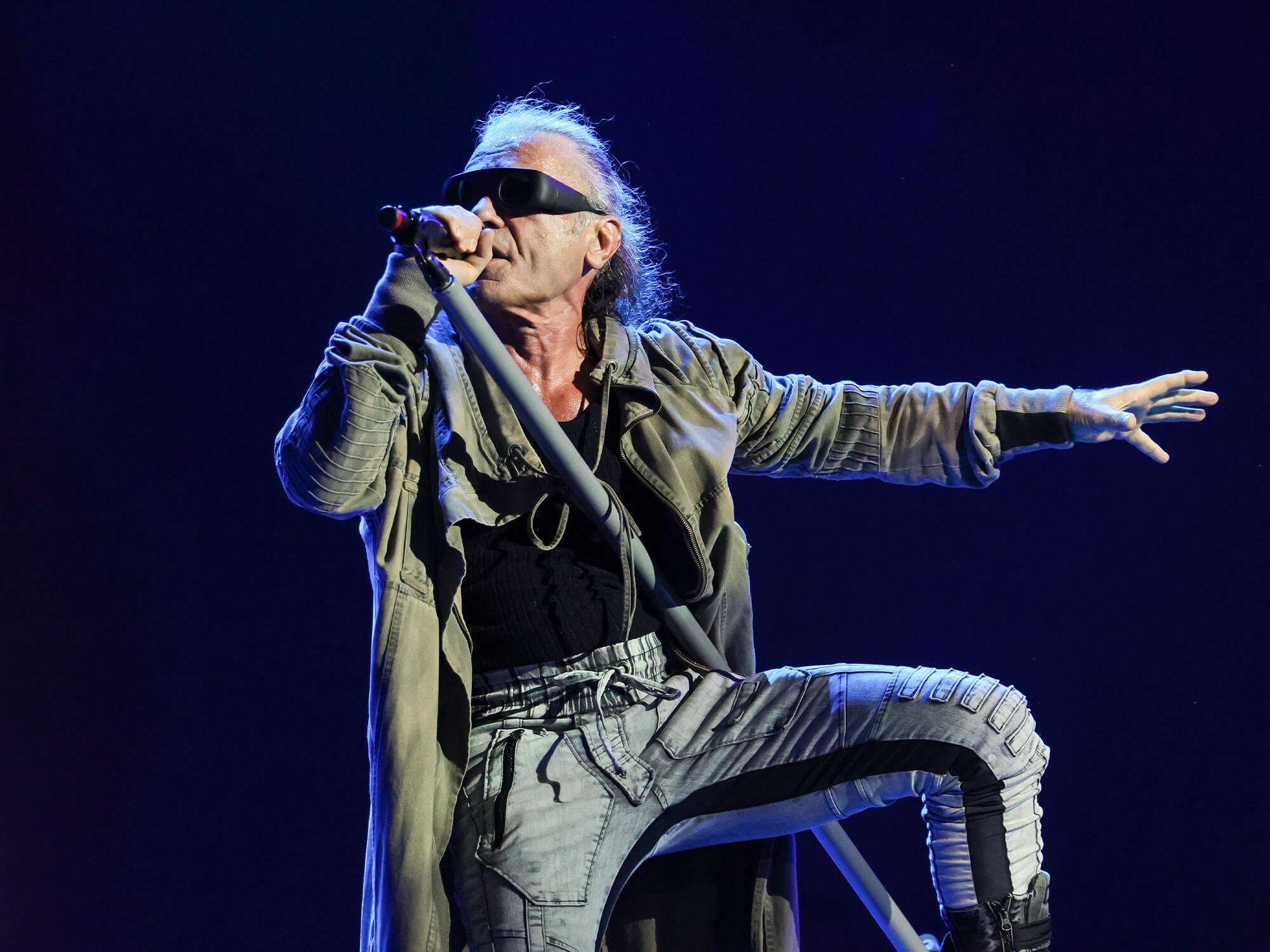 Singer in a green coat and dark visor-style glasses sings into a microphone on a blue-lit stage